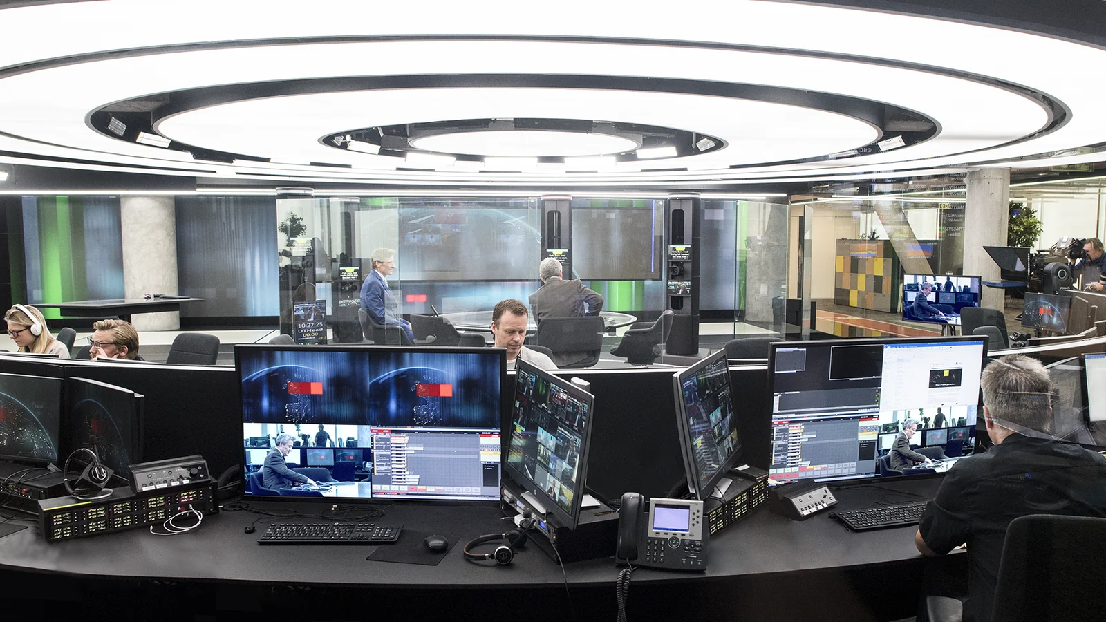A large broadcast gallery under a circular ring light, with several operators seated along a curved desk in front of stacked monitors and production keypads