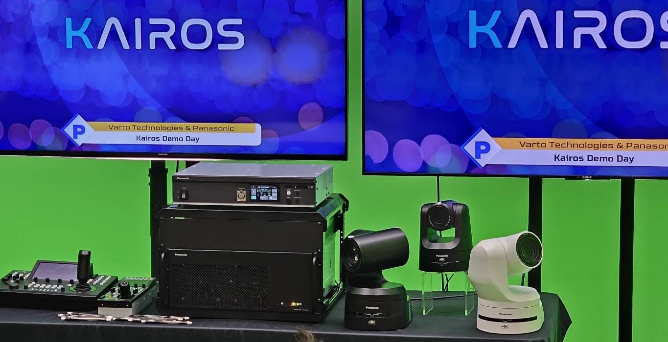 Three white PTZ cameras, a controller and a rack mainframe on a green-screen bench, with the Kairos wordmark on two monitors behind