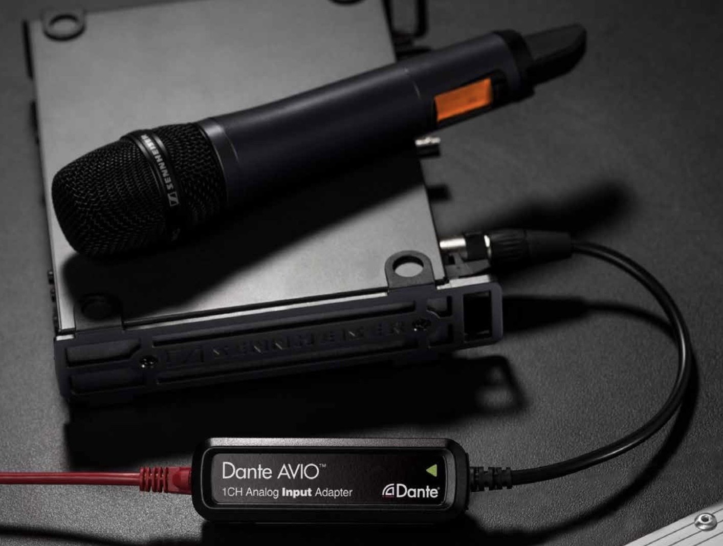 A Dante AVIO one-channel analog input adapter plugged into the XLR output of a wireless microphone receiver, with the handheld microphone resting on the receiver