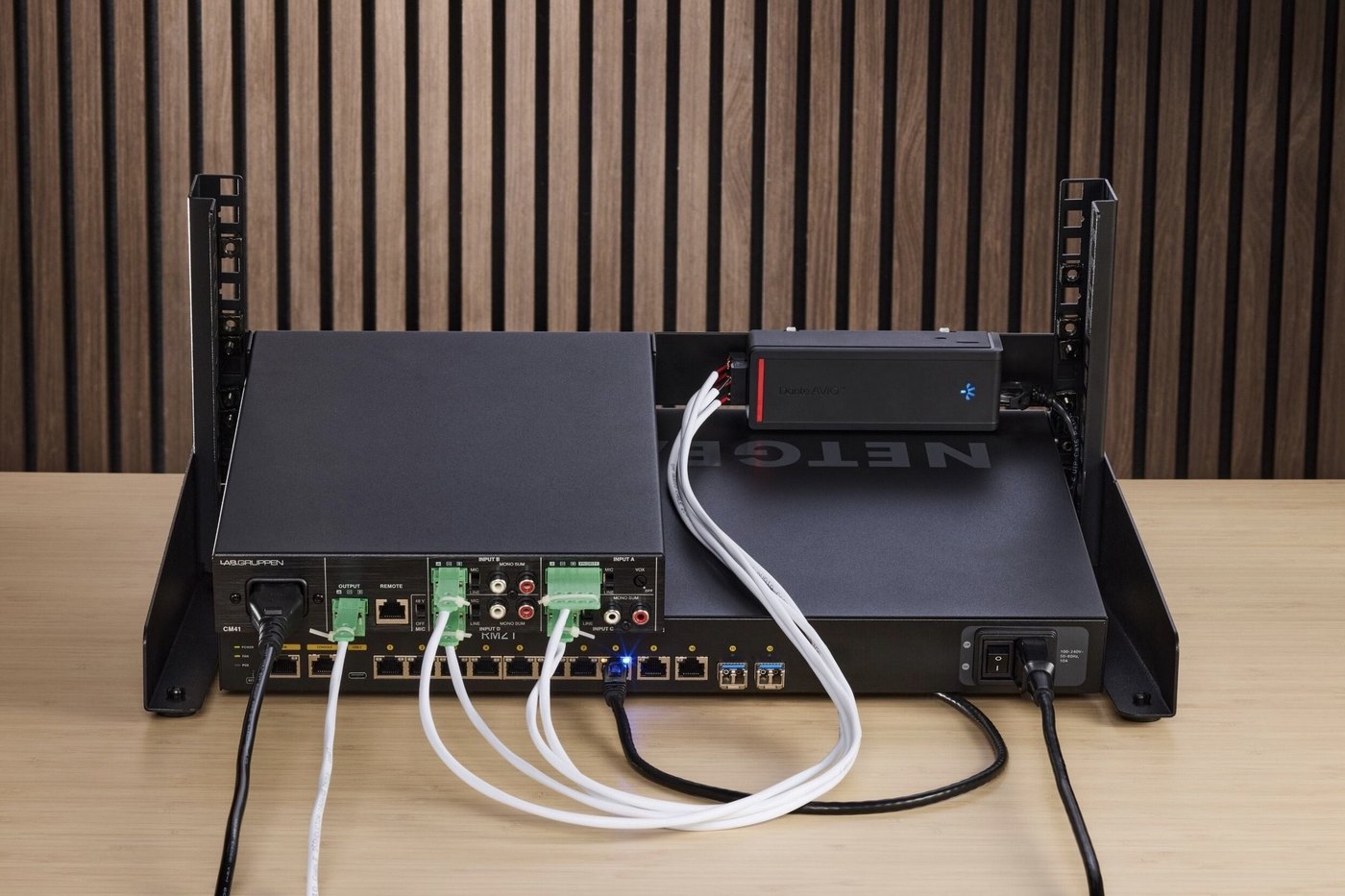 A Dante AVIO Install adapter fixed to a rack shelf and wired by Euroblock to an amplifier, beside a network switch