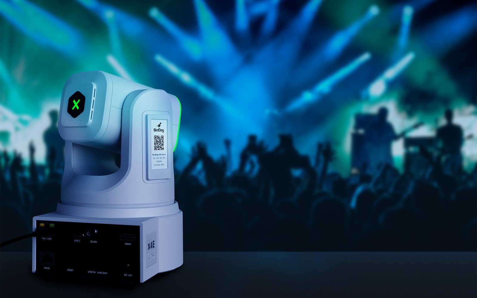 A white BirdDog X4E Ultra PTZ camera seen from the rear, with its connector panel, in front of a lit concert stage