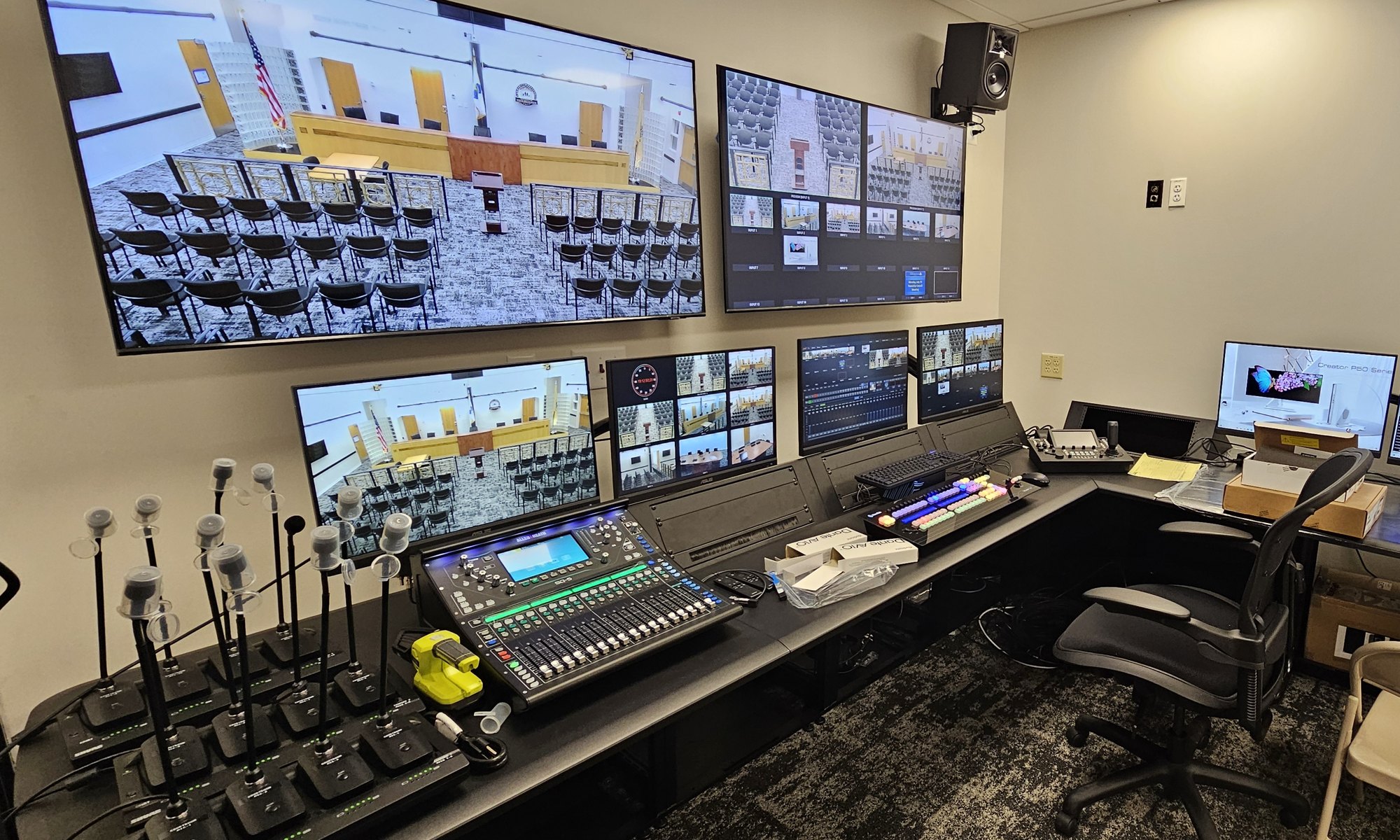 Township control room with wall monitors showing council chamber camera views, an audio console, a switcher panel and wireless microphone chargers