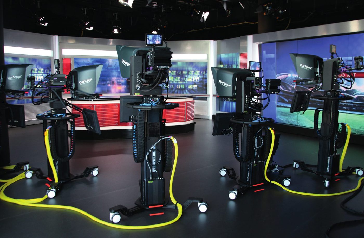 Three pedestal mounted studio cameras, each carrying a CueScript prompter, on a dark studio floor with video walls behind