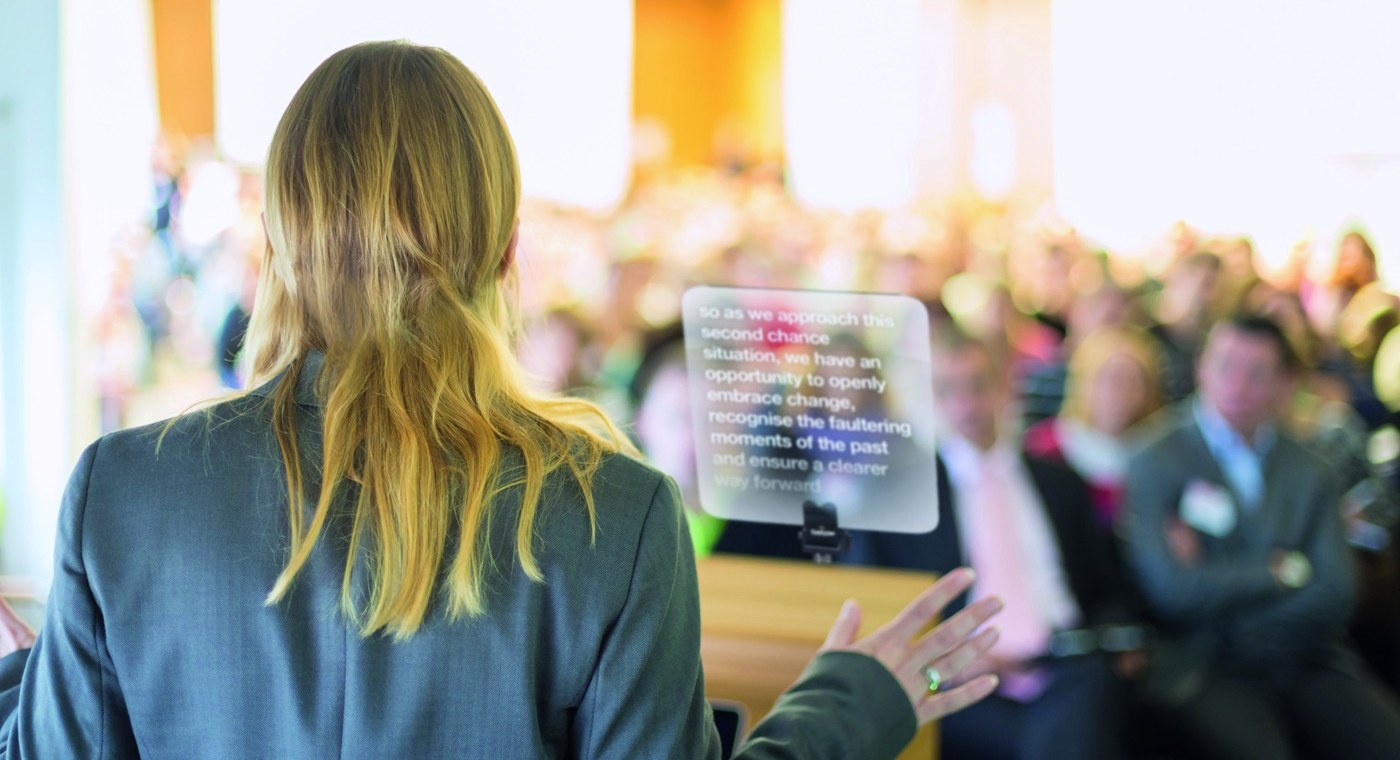 A presenter at a lectern reading from a CueScript glass speech prompter in front of an audience
