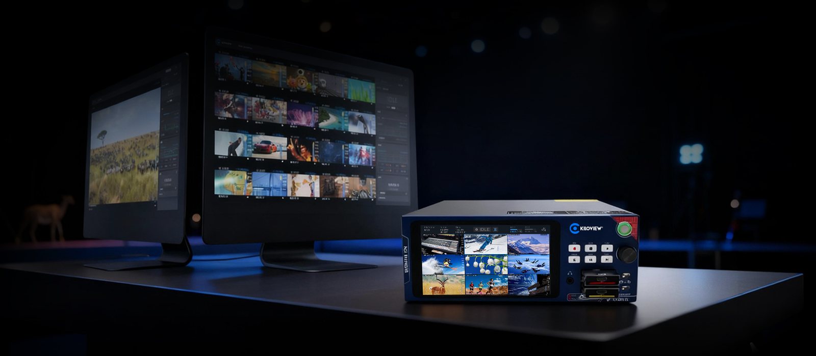Kiloview CUBE R1 recorder on a desk, its front screen showing a grid of recorded channels, with editing monitors behind