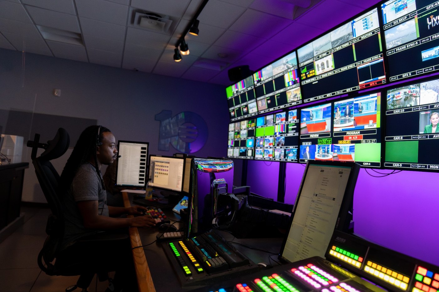 Operator at a production control position with a Ross Video switcher panel and control screens beneath a lit monitor wall