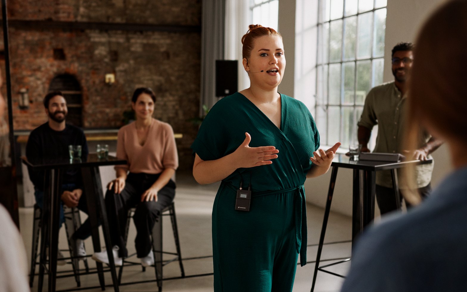 A presenter speaking with a headset microphone and a clipped wireless bodypack to a small audience at high tables