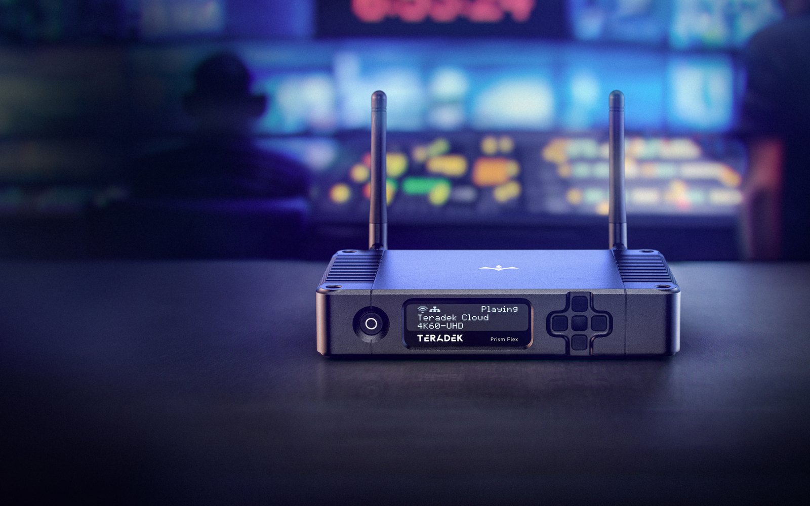 A Teradek Prism Flex encoder on a dark desk in front of a blurred control room and monitor wall