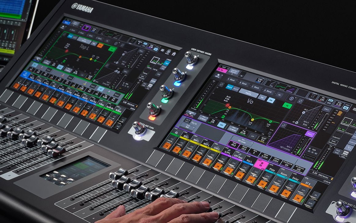 An operator's hand on the faders of a Yamaha DM5 console, with its two touchscreens lit
