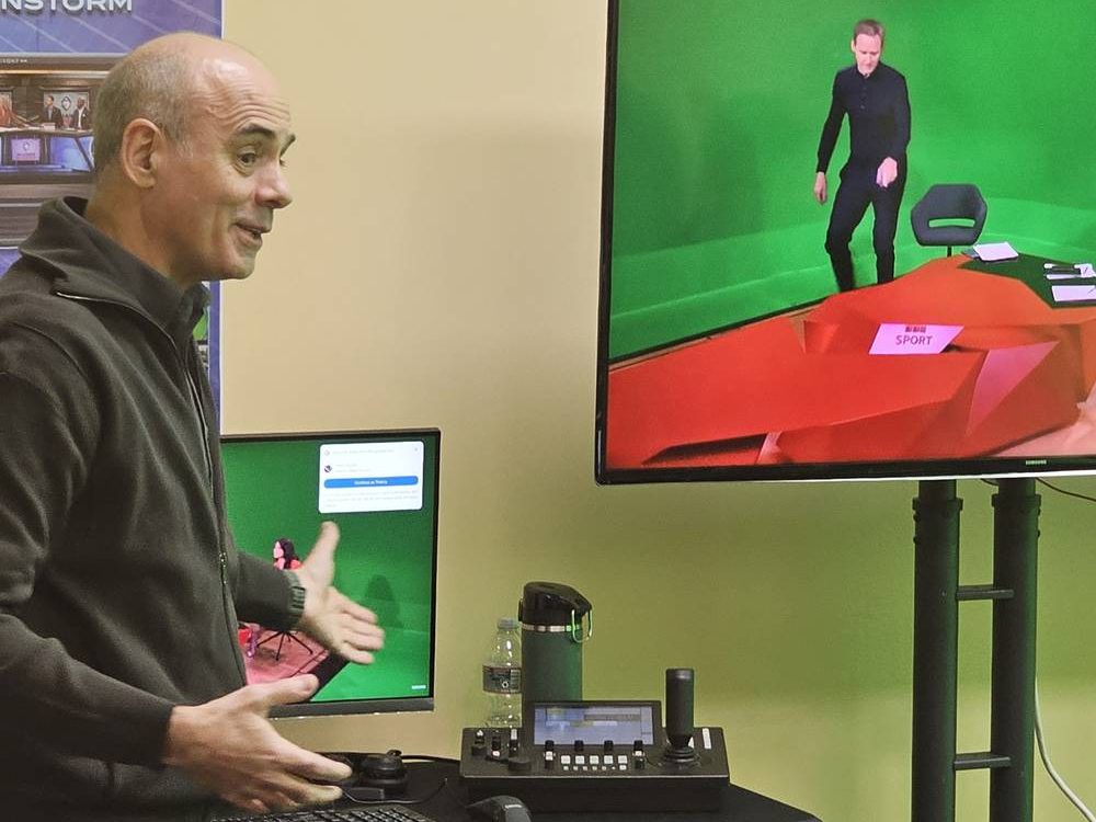 A presenter driving a keyed shot on a monitor beside the green cyclorama