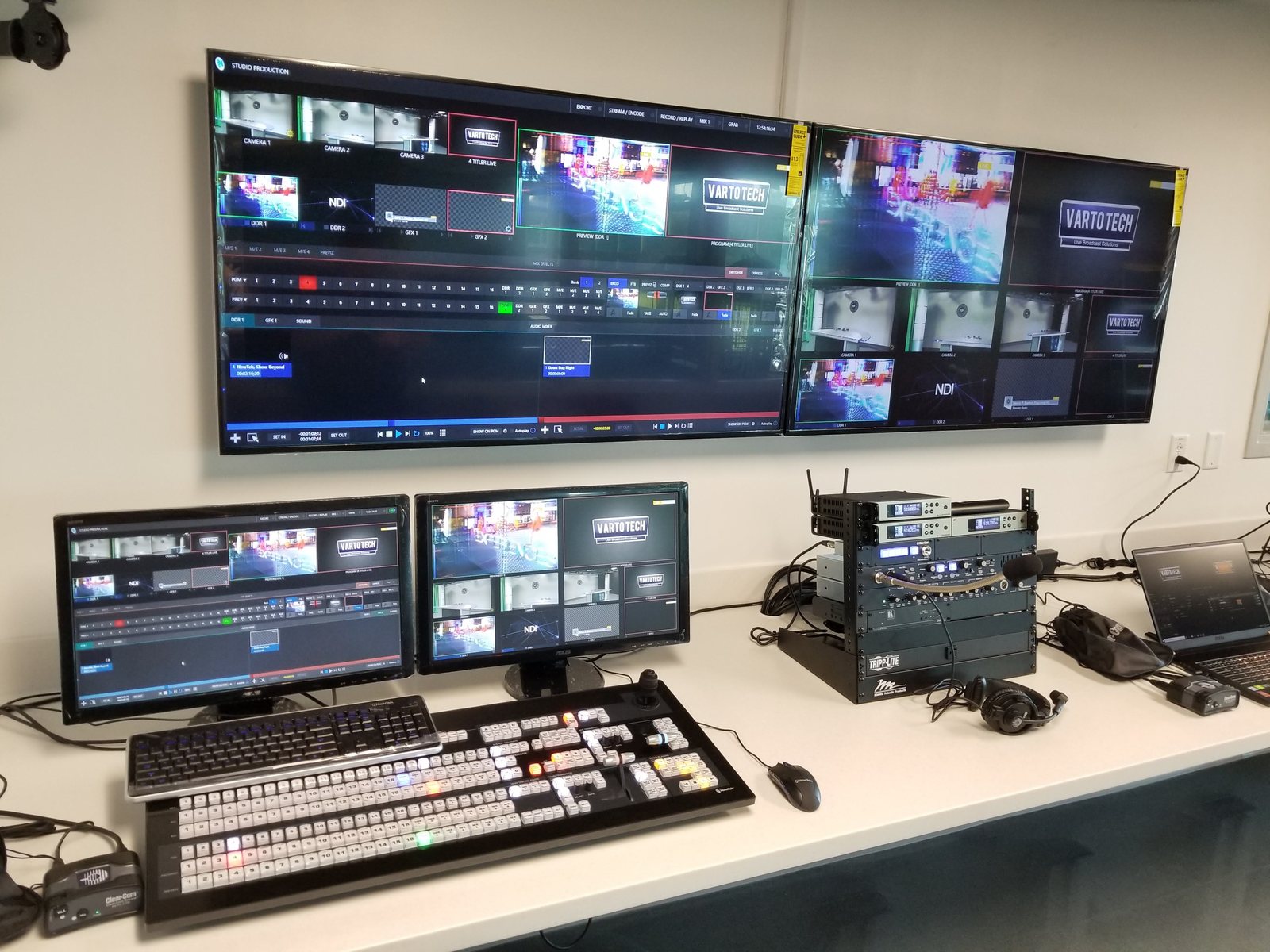 A school control room: a TriCaster interface and multiviewer on two wall monitors, a hardware control surface and two desk monitors below, and a small equipment rack