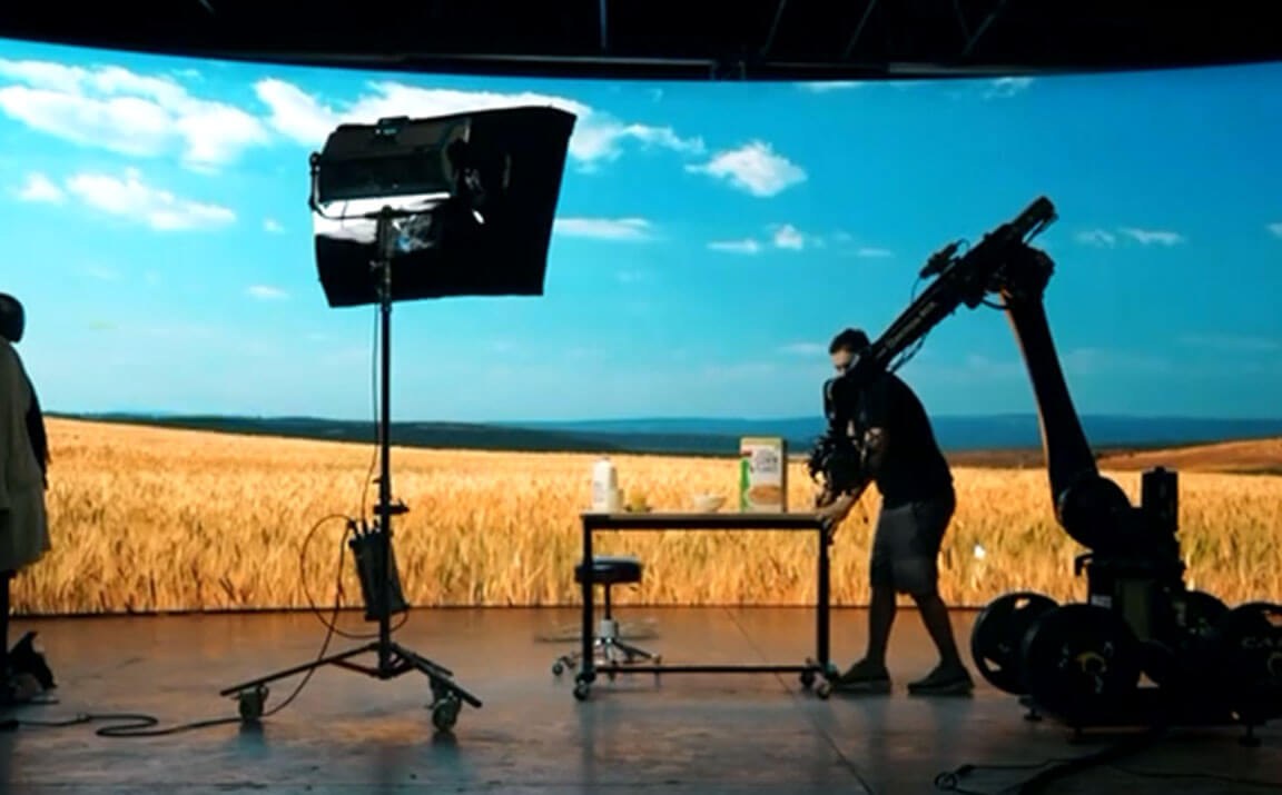 Silhouetted crew film in front of an LED wall showing a wheat field under a blue sky, with a robotic camera arm and a softbox light