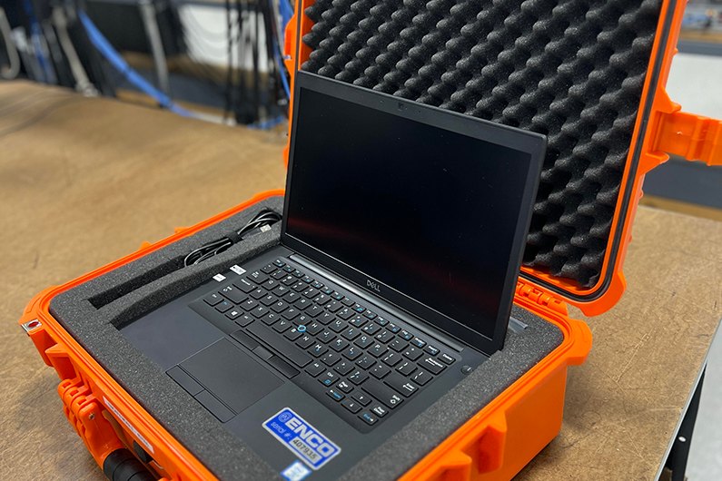 A laptop with an ENCO asset label sitting open in an orange hard-shell carrying case on a workbench