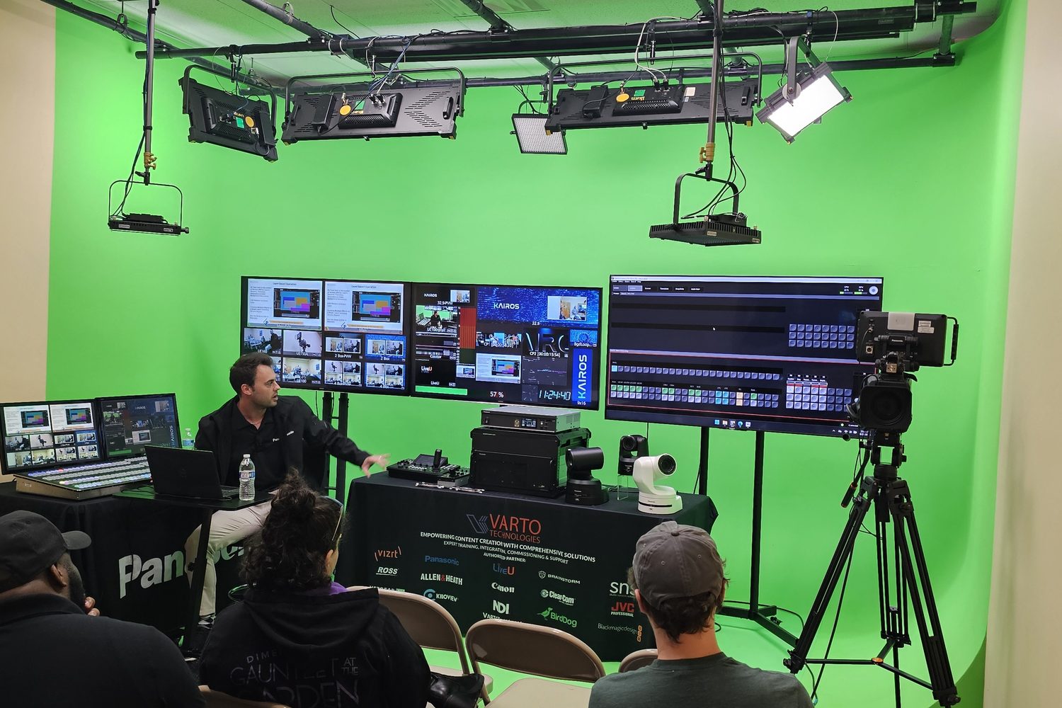 Attendees in folding chairs watch a KAIROS demonstration in a green-screen studio, with multiviewer monitors, a studio camera on a tripod and PTZ cameras on the table