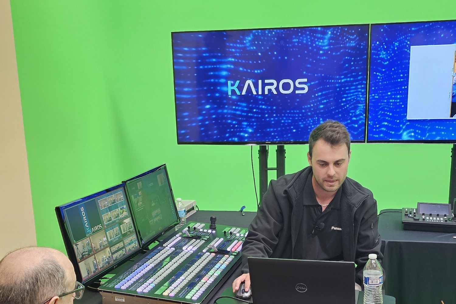 A presenter at a laptop beside a lit KAIROS control panel and two monitors, with the KAIROS logo on a large screen behind him