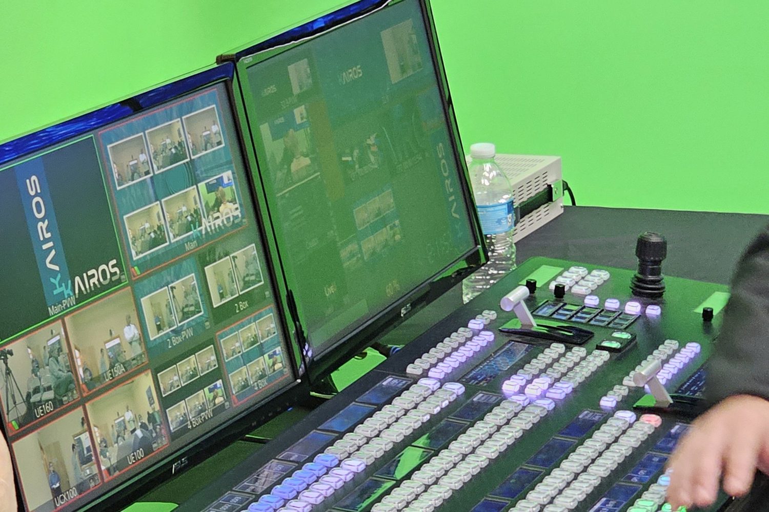 A KAIROS control panel with rows of lit buttons and a T-bar beside two monitors showing a KAIROS multiviewer of camera tiles