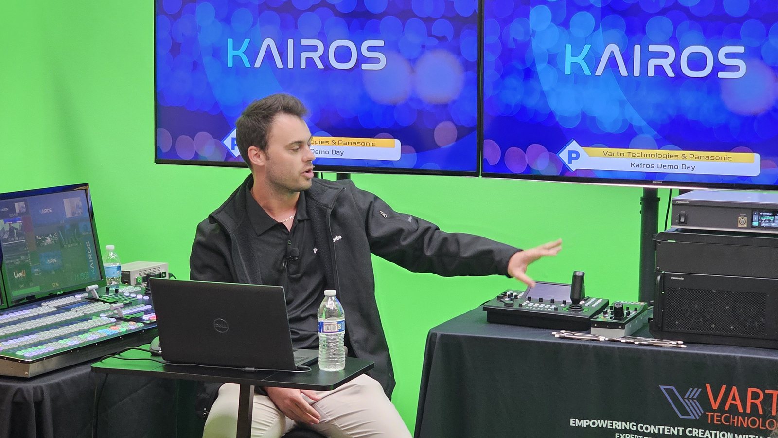 A presenter in a green-screen studio gestures toward a camera controller and a rack mainframe beside a large switcher panel, with KAIROS demo day graphics on the screens behind