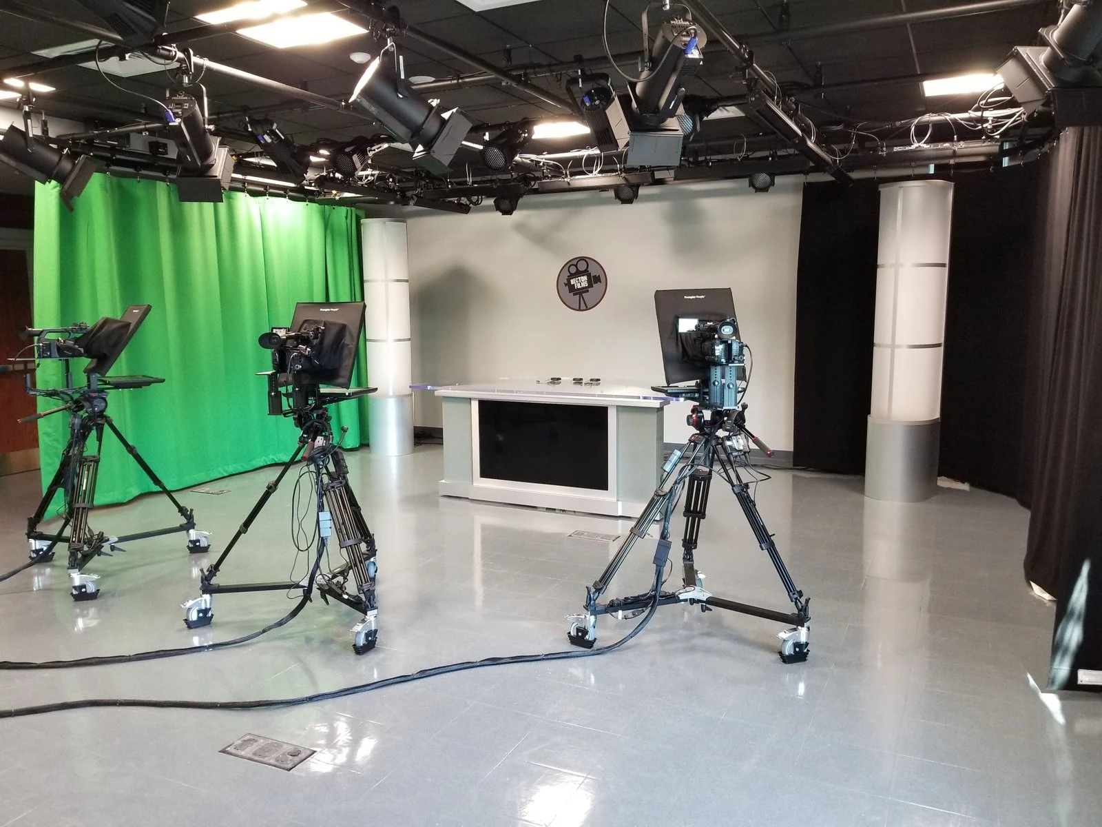 A school television studio with three prompter-equipped cameras, an anchor desk and a green screen under a lighting grid