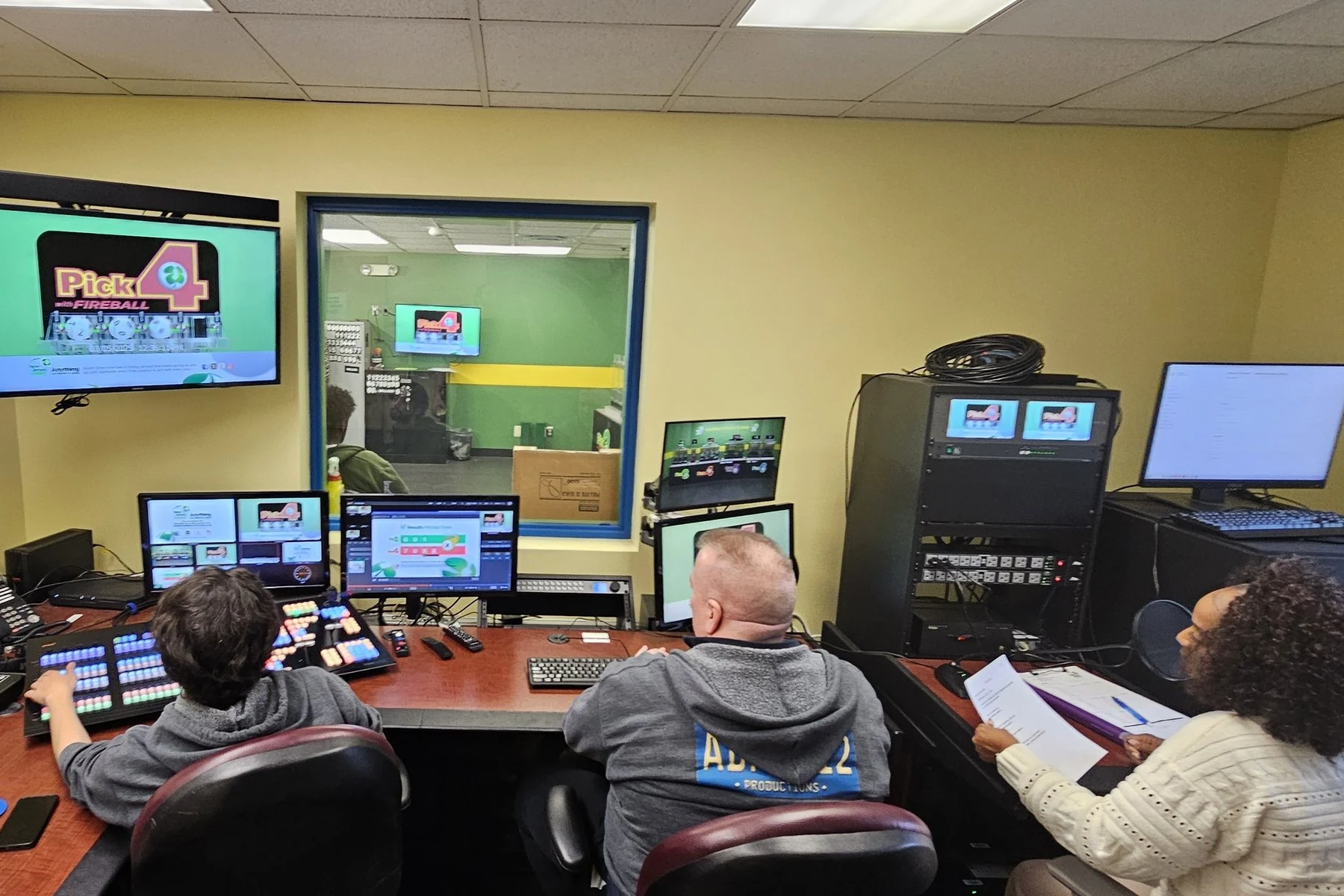 New Jersey Lottery control room in March 2025, three people at the switcher and audio positions with a Pick 4 draw graphic on the wall monitor