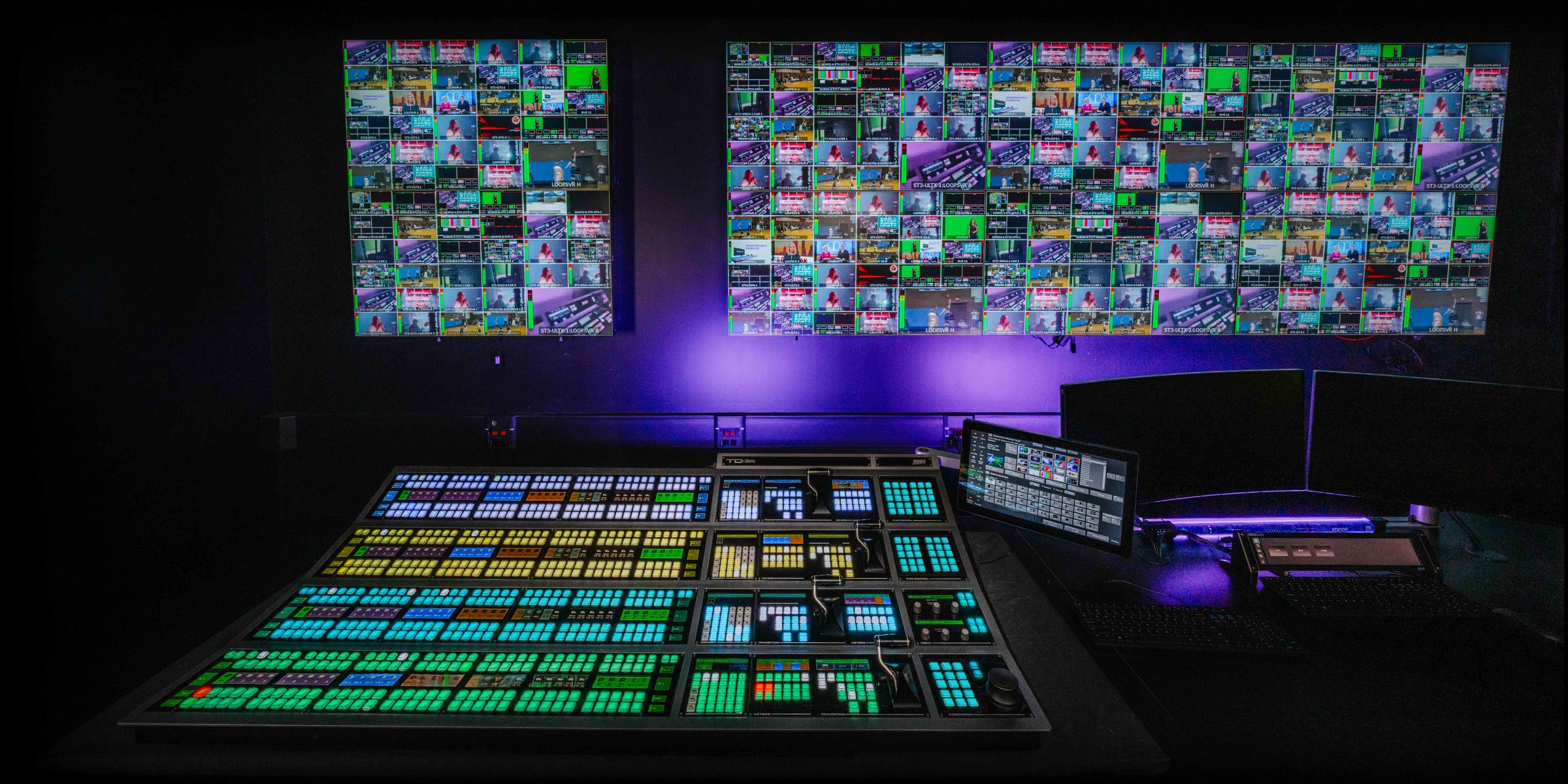 A four-row TouchDrive switcher panel glowing in a dark control room, below two walls of multiviewer tiles lit in purple