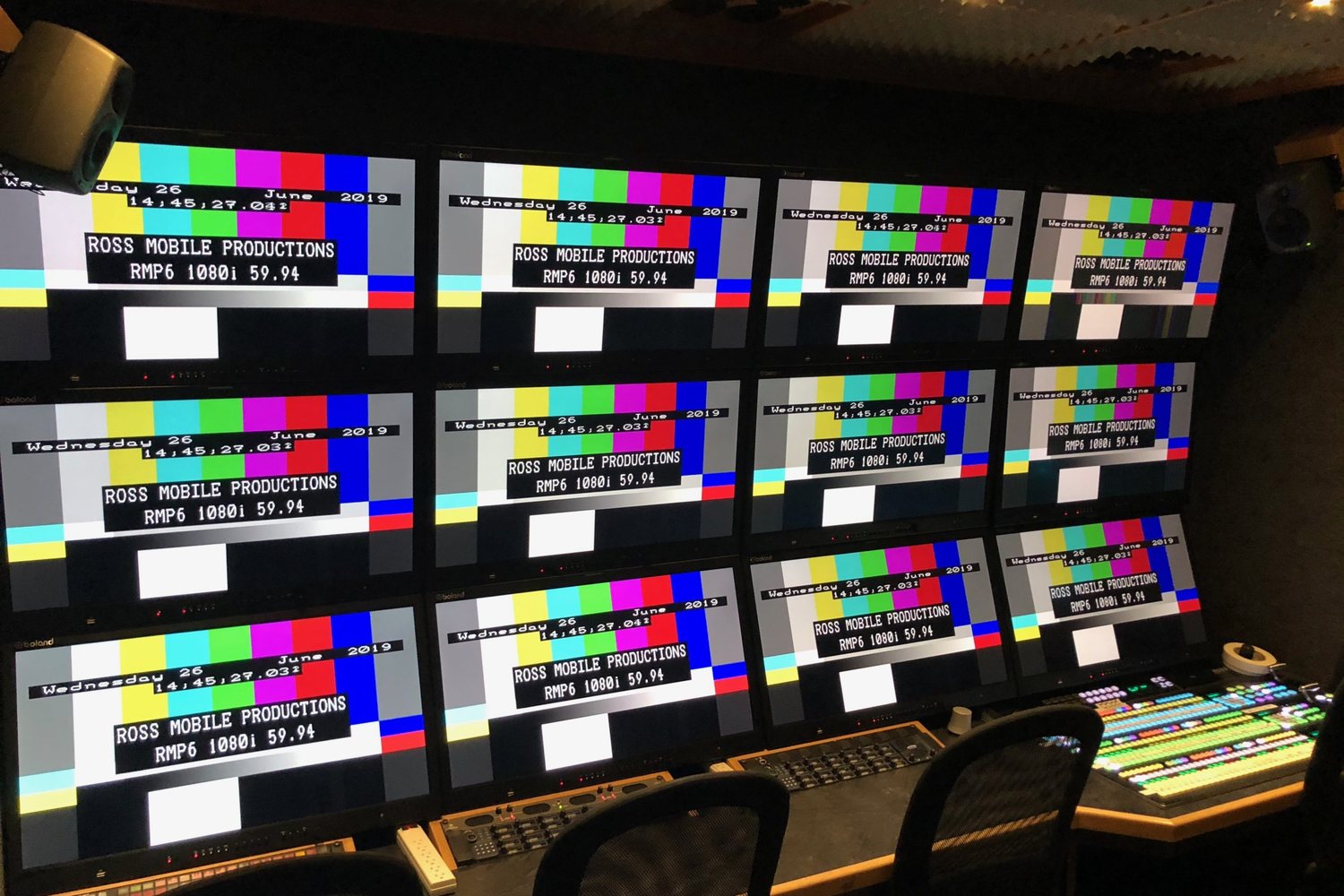 A production truck control room with three rows of monitors showing color bars above a lit switcher panel